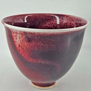 May include: A handmade ceramic bowl with a deep burgundy glaze. The bowl has a wide opening, a rounded body, and a small, circular base. The interior and exterior feature a gradient of colors, transitioning from a lighter red to a darker, almost black hue.