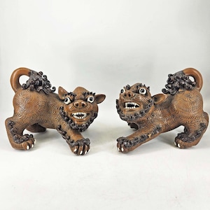 May include: A pair of brown ceramic foo dogs with intricate details. The foo dogs are in a playful pose, with one paw raised and the other on the ground. They have a fierce expression with open mouths and sharp teeth.