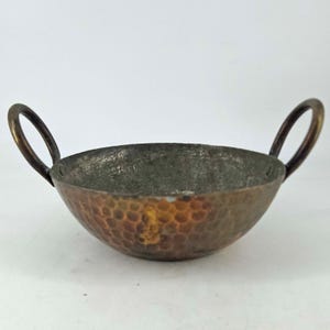 May include: A small, hammered copper bowl with two arched handles. The bowl has a textured surface and a tarnished appearance, with a mix of copper and darker tones. The handles are a darker, aged copper color.