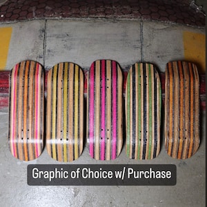 Op de afbeelding: Vijf fingerboards met verschillende grafische ontwerpen. De fingerboards zijn gemaakt van hout en hebben zwarte, gele, roze, groene en oranje strepen. De tekst "Graphic of Choice w/ Purchase" staat onderaan de afbeelding.