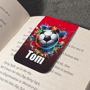 May include: A red, black, and white bookmark with a soccer ball graphic and the name "Tom" printed on it. The soccer ball is surrounded by colorful paint splatters.