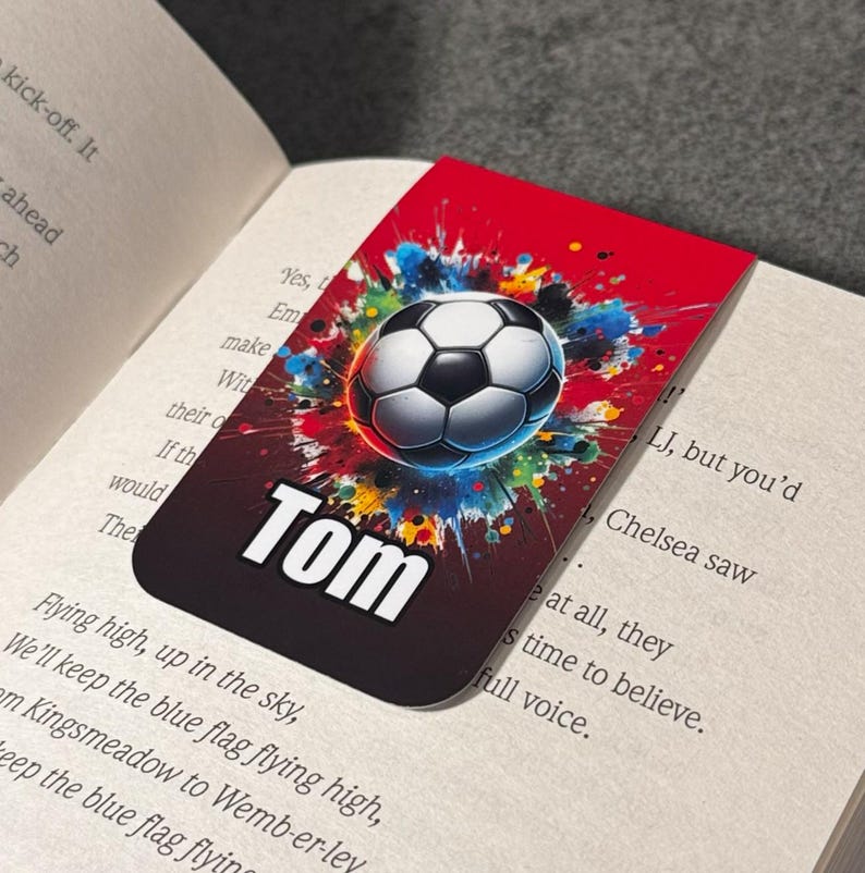 May include: A red, black, and white bookmark with a soccer ball graphic and the name "Tom" printed on it. The soccer ball is surrounded by colorful paint splatters.