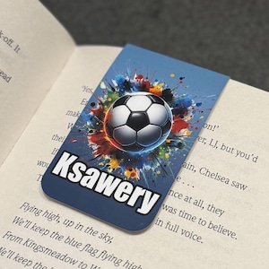 May include: A blue bookmark with a colorful paint splatter design featuring a soccer ball in the center. The name "Ksawery" is printed in white letters at the bottom of the bookmark.