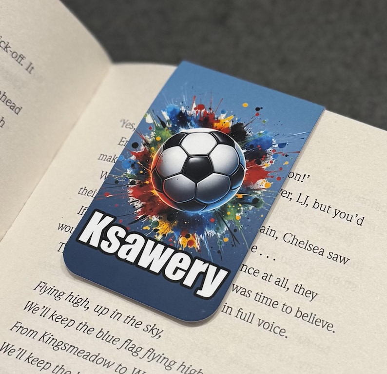 May include: A blue bookmark with a colorful paint splatter design featuring a soccer ball in the center. The name "Ksawery" is printed in white letters at the bottom of the bookmark.