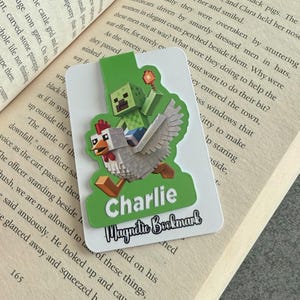 May include: A rectangular magnetic bookmark with a pixelated green zombie riding a white chicken. The bookmark has the name "Charlie" and the text "Magnetic Bookmark" on a green background. The bookmark is placed on an open book.