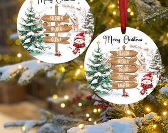 Può includere: Due decorazioni natalizie rotonde con un design festoso. Ogni ornamento presenta una scena invernale con un pupazzo di neve, un albero di Natale e un cartello stradale con nomi. Il testo "Merry Christmas" è scritto in alto. Nastro rosso per appendere.