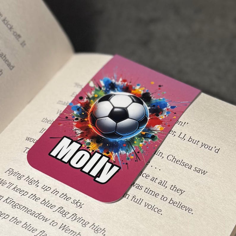 May include: Pink bookmark with a colorful paint splatter design featuring a soccer ball in the center. The name "Molly" is printed in white at the bottom of the bookmark.