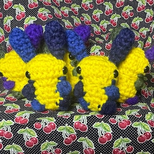 Puede incluir: Un grupo de pequeñas criaturas tejidas a ganchillo a mano con cuerpos de color amarillo brillante, orejas azul oscuro y detalles en azul oscuro. Cada criatura tiene dos pequeños ojos de cuentas negras. Las figuras están agrupadas.