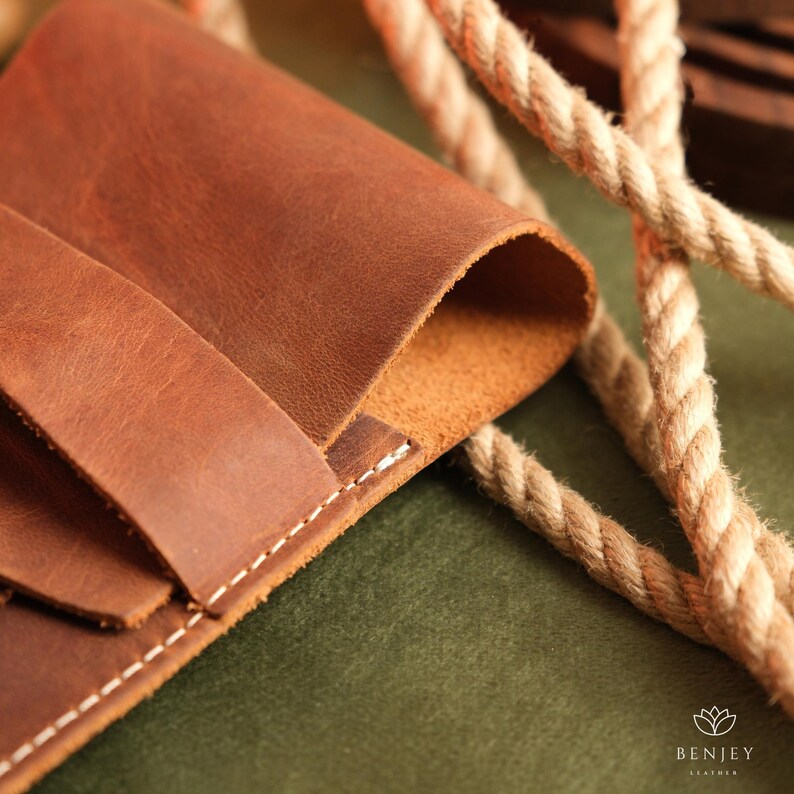 May include: Close-up of a brown leather pouch with stitching detail. The pouch is folded over and has a flap with a stitched edge. The pouch is on a green surface with a rope.