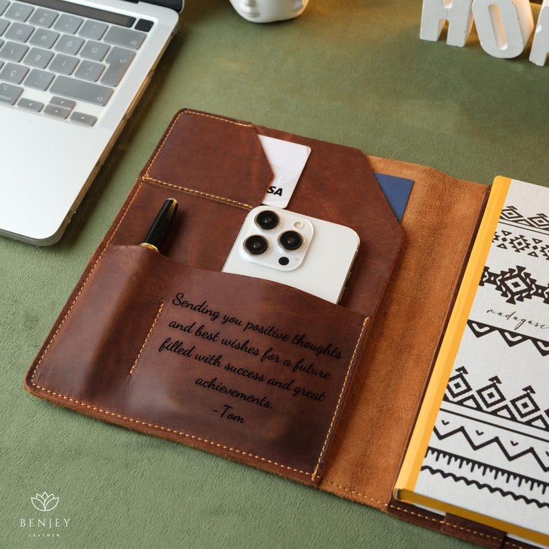 Może przedstawiać: Brązowy sk&oacute;rzany notes z telefonem i długopisem w środku. Notes zawiera spersonalizowany napis: "Sending you positive thoughts and best wishes for a future filled with success and great achievements. -Tom".