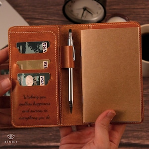 May include: Open leather notebook with a pen, credit card slots, and a notepad. The notebook is tan with a stitched border. The text "Wishing you endless happiness and success in everything you do" is visible.