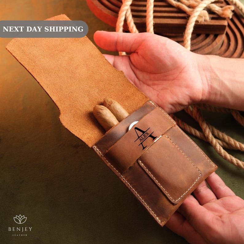 May include: A brown leather cigar case with a personalised name, "Alex", engraved on the front. The case has a flap that opens to reveal two cigars inside.