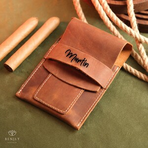 May include: A brown leather cigar case with a flap that has the name "Martin" embossed on it. Two cigars are visible in the background.