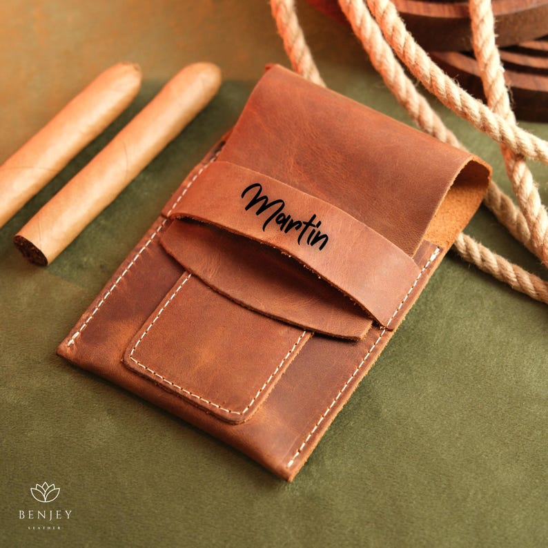 May include: A brown leather cigar case with a flap that has the name "Martin" embossed on it. Two cigars are visible in the background.
