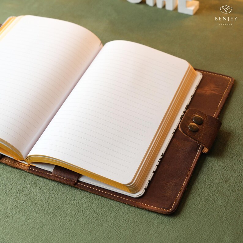 Może przedstawiać: Brązowy, sk&oacute;rzany notes z pozłacanymi krawędziami stron jest otwarty, ukazując liniowany papier. Notes ma zapięcie na klapkę z metalowym guzikiem.