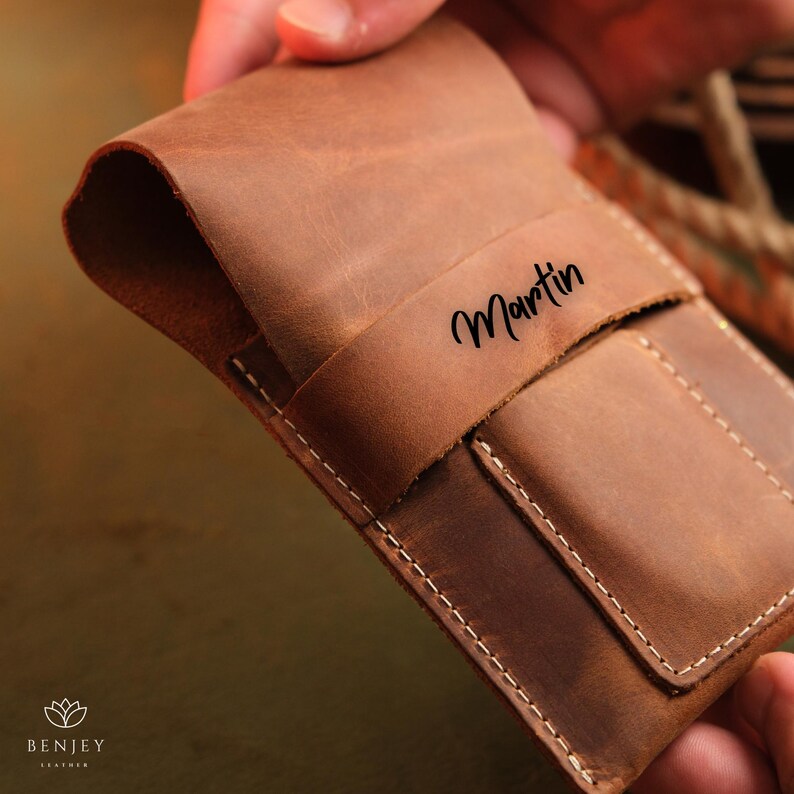 May include: A brown leather wallet with a flap closure and a stitched pocket. The wallet is personalised with the name "Martin" in black lettering.