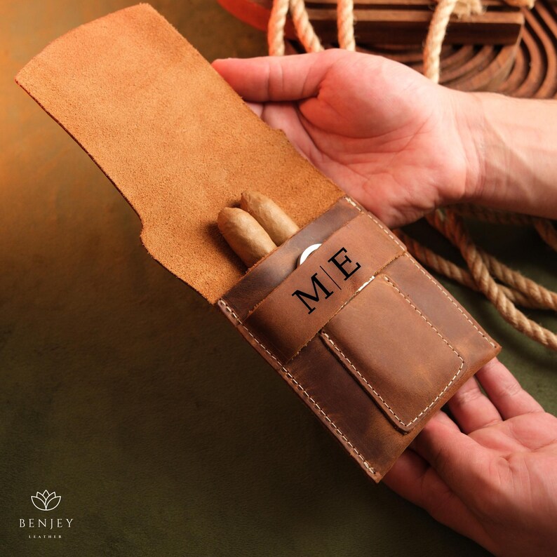 May include: A brown leather cigar case with two cigars inside. The case has a pocket for a cigar cutter and is personalised with the initials "M/E".