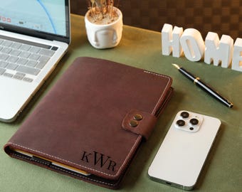 May include: A brown leather bound journal with a flap closure and a personalized monogram "KWR" on the front. The journal is on a green surface with a white phone and a pen.