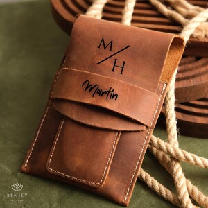 May include: A brown leather phone case with a flap closure. The case is personalised with the initials "M/H" and the name "Martin".