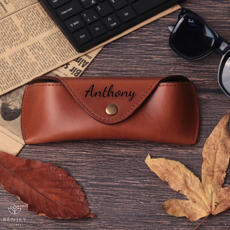 Pode incluir: Um estojo de &oacute;culos de couro castanho com o nome "Anthony" gravado na frente. O estojo &eacute; fechado com um fecho de press&atilde;o cor de lat&atilde;o. Um par de &oacute;culos de sol pretos e um teclado est&atilde;o no fundo.