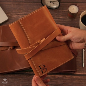 May include: A tan leather journal with a strap closure. The journal has the name "Dylan" and the letter "D" engraved on the front. The journal is shown open, revealing the interior pages. The stitching is white.