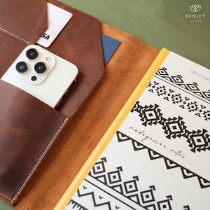 May include: A brown leather notebook cover with a white smartphone and a credit card in the pockets. A patterned notebook with the text "madagascar notes" is visible. The leather has a distressed look.