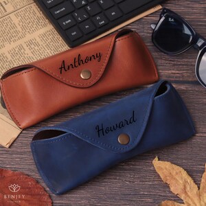 Pode incluir: Duas capas de &oacute;culos de couro, uma castanha e outra azul, com nomes personalizados. A capa castanha tem a inscri&ccedil;&atilde;o "Anthony", enquanto a capa azul tem a inscri&ccedil;&atilde;o "Howard". Cada capa tem um fecho de press&atilde;o. &Oacute;culos de sol e teclado no fundo.