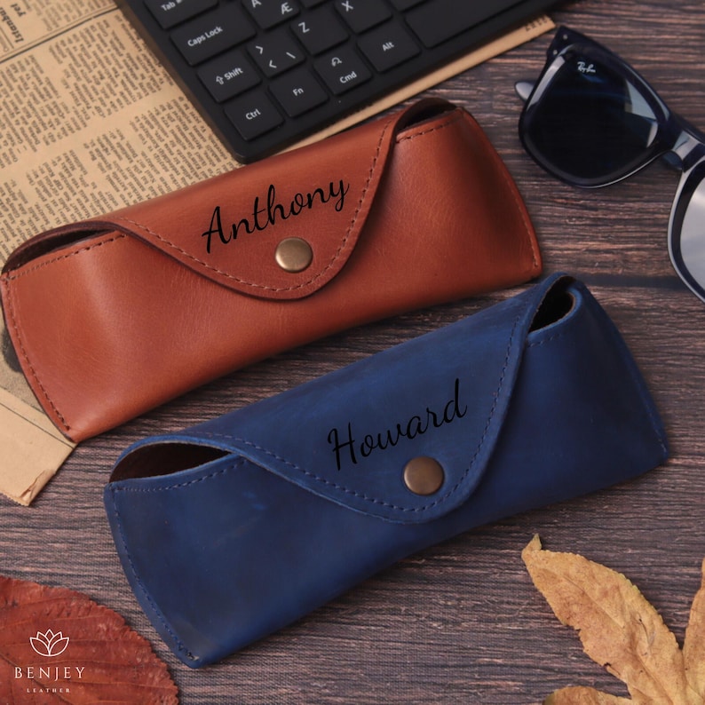 Pode incluir: Duas capas de &oacute;culos de couro, uma castanha e outra azul, com nomes personalizados. A capa castanha tem a inscri&ccedil;&atilde;o "Anthony", enquanto a capa azul tem a inscri&ccedil;&atilde;o "Howard". Cada capa tem um fecho de press&atilde;o. &Oacute;culos de sol e teclado no fundo.