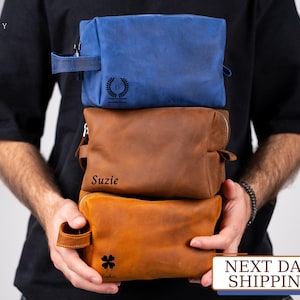 May include: Three leather toiletry bags stacked on top of each other. The top bag is blue and has a monogram with the letter D and a laurel wreath. The middle bag is brown and has the name "Suzie" printed on it. The bottom bag is brown and has a four-leaf clover graphic and the word "Sophia" printed on it.