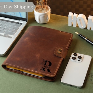 May include: A brown leather journal with a personalized initial and name is displayed open on a green surface. A white smartphone, a pen, and a laptop are also present. The words "Next Day Shipping" are in the top left corner.