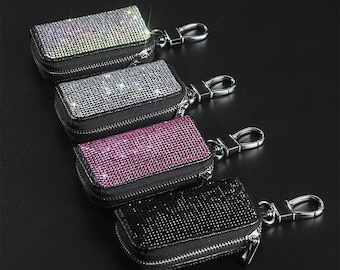 May include: Four rectangular keychains with sparkling rhinestone embellishments in silver, gray, pink, and black. Each keychain has a black zipper and a silver-colored clasp. The keychains are displayed on a dark surface.
