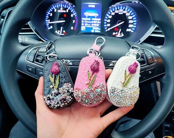 May include: Three keychains in black, pink, and white, each adorned with a jeweled tulip design. The keychains have a textured finish and are embellished with sparkling crystals. The background shows a car's steering wheel and dashboard.