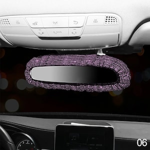 May include: A car rearview mirror with a purple rhinestone cover. The cover is made of small, sparkling rhinestones that completely encase the mirror. The mirror is mounted on the car's ceiling, with various control buttons visible above.