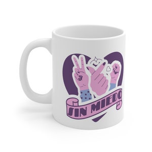 Könnte beinhalten: Weiße Keramik-Tasse mit einem lila Herz-Design. Das Design beinhaltet Handgesten und den Text "SIN MIEDO" auf einem Banner. Die Hände sind rosa mit blau-weiß gemusterten Ärmeln.
