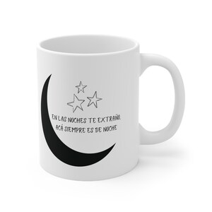 May include: White ceramic mug featuring a black crescent moon and three stars. The mug has the text "EN LAS NOCHES TE EXTRAÑO. ACÁ SIEMPRE ES DE NOCHE" in black. The mug has a curved handle.