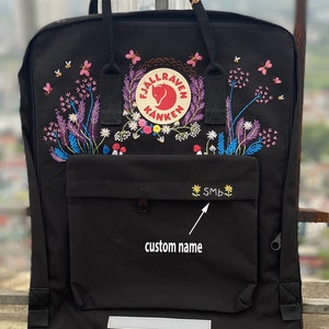 May include: A black backpack featuring a Fjällräven Kånken logo patch surrounded by colorful floral and insect embroidery. The front pocket displays the embroidered text SMB flanked by two small yellow flowers, with a reflective strip at the base.