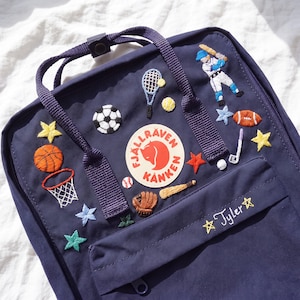 Könnte beinhalten: Ein marineblauer Rucksack mit einem runden Fjällräven Kånken-Logoaufnäher und verschiedenen gestickten Sportmotiven, darunter ein Basketball, ein Fußball, ein Tennisschläger, ein Baseballspieler und ein Golfschläger. Der Name Tyler ist auf der Tasche.