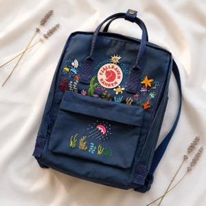 May include: Navy blue backpack with embroidered ocean-themed designs, including jellyfish, coral, and seaweed. The backpack features a front pocket and top handle. The Fjallraven Kanken logo is visible on the front.