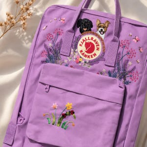 Può includere: Uno zaino lavanda con ricami floreali e di cani. Lo zaino presenta una tasca frontale, una maniglia superiore e il logo Fjallraven. Il ricamo include fiori rosa, api, un cane nero e un cane marrone.