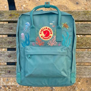 Könnte beinhalten: Türkisfarbener Rucksack mit gestickten Meeresmotiven, darunter Quallen, Korallen und eine Schildkröte. Mit einer Vordertasche und einem oberen Griff. Der Rucksack hat das Fjallraven Kanken Logo.