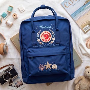 May include: Navy blue backpack with embroidered sea-themed designs, including starfish, seashells, and coral. The backpack has a top handle and a front pocket. The word "Marina" is embroidered above the logo. Other items include a camera, headphones, and a teddy bear.