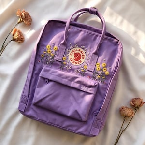 Peut inclure: Un sac à dos lavande avec des broderies de tournesols jaunes et un logo de renard rouge. Le sac à dos a une poche avant et une poignée supérieure. Le mot "Angelica" est brodé sur la poche avant.