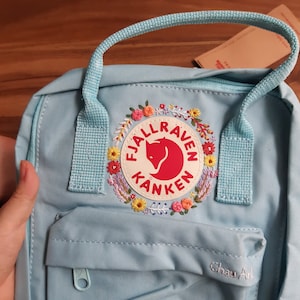 Puede incluir: Una mochila azul claro con un parche floral bordado con el texto "Fjallraven Kanken" y un logotipo de zorro rojo. La mochila tiene un asa superior, un bolsillo frontal y cierres de cremallera. El texto "Chau Art" está bordado en el bolsillo frontal.