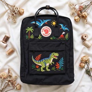 Könnte beinhalten: Ein schwarzer Rucksack mit gestickten prähistorischen Motiven, darunter ein grüner Dinosaurier auf der Vordertasche, zwei blaue Flugsaurier, eine Palme, ein Vulkan und eine gelbe Sonne, mit einem runden Fjällräven Kånken Logo oben mittig.