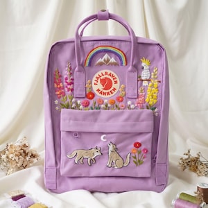May include: A light purple backpack featuring embroidered designs including a rainbow over mountains, a white cockatoo, colorful wildflowers, and a front pocket with two wolves howling at a crescent moon. The Fjällräven Kånken logo is centered.