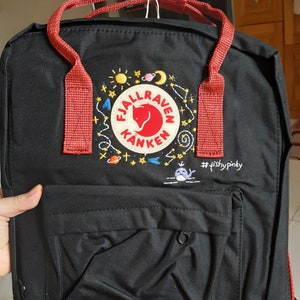 May include: Black backpack with red straps and handles. The front pocket has a small, round zipper. The backpack features an embroidered logo with a red fox, sun, moon, and stars. The text "#fishypinky" is also embroidered on the backpack.