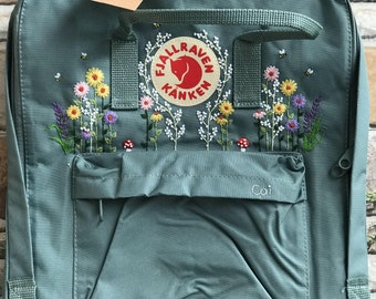 May include: A teal backpack with a floral embroidery design featuring daisies, lavender, and mushrooms. The backpack has a front pocket with the name "Cai" embroidered on it. The brand logo "Fjallraven Kanken" is visible on the front.