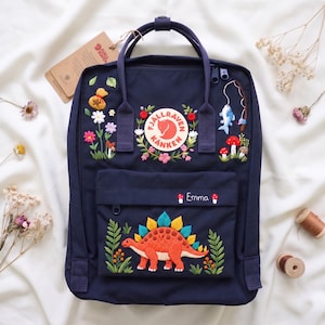 Op de afbeelding: Een marineblauwe Fjällräven Kånken rugzak met aangepast borduurwerk van een dinosaurus, bloemen, paddenstoelen en een visscène. Het voorvak toont de naam Emma in witte tekst, geflankeerd door twee kleine rood-witte paddenstoelmotieven.