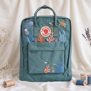 Può includere: Uno zaino Fjällräven Kånken color verde acqua con ricamo personalizzato di una scena sottomarina con coralli, alghe, una tartaruga marina e meduse. La tasca frontale mostra il nome Maria ricamato in corsivo sotto dei pesciolini.