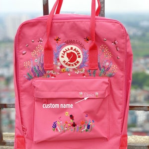 May include: A pink backpack featuring a circular Fjällräven Kånken logo patch, embroidered floral and insect motifs, and a front pocket with the embroidered name Elexia and the text custom name with an arrow pointing to the name.